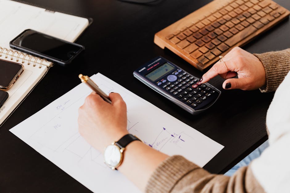 Home Close-up of hands working with a calculator and notebook on a desk, analyzing documents.